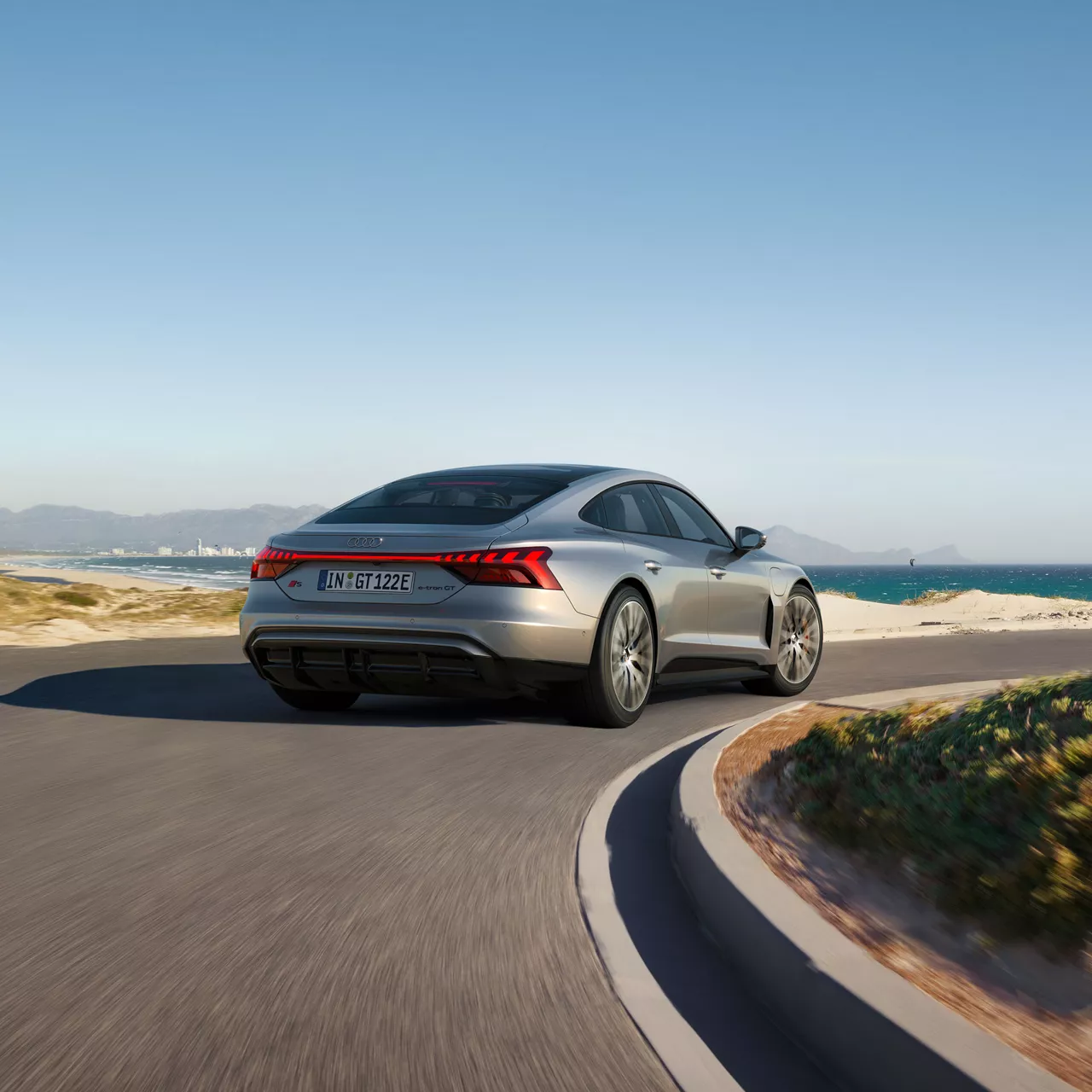 Ein silberner Audi S e-tron GT fährt auf einer kurvigen Küstenstraße mit Blick auf das Meer und eine Stadt im Hintergrund.