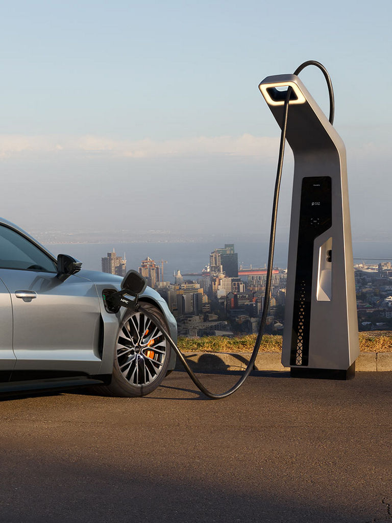 A silver Audi S e-tron GT is charging at a charging station with a view of a city. A man in a suit walks alongside the car.