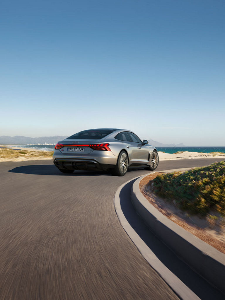  A silver Audi S e-tron GT drives along a winding coastal road with a view of the sea and a city in the background.