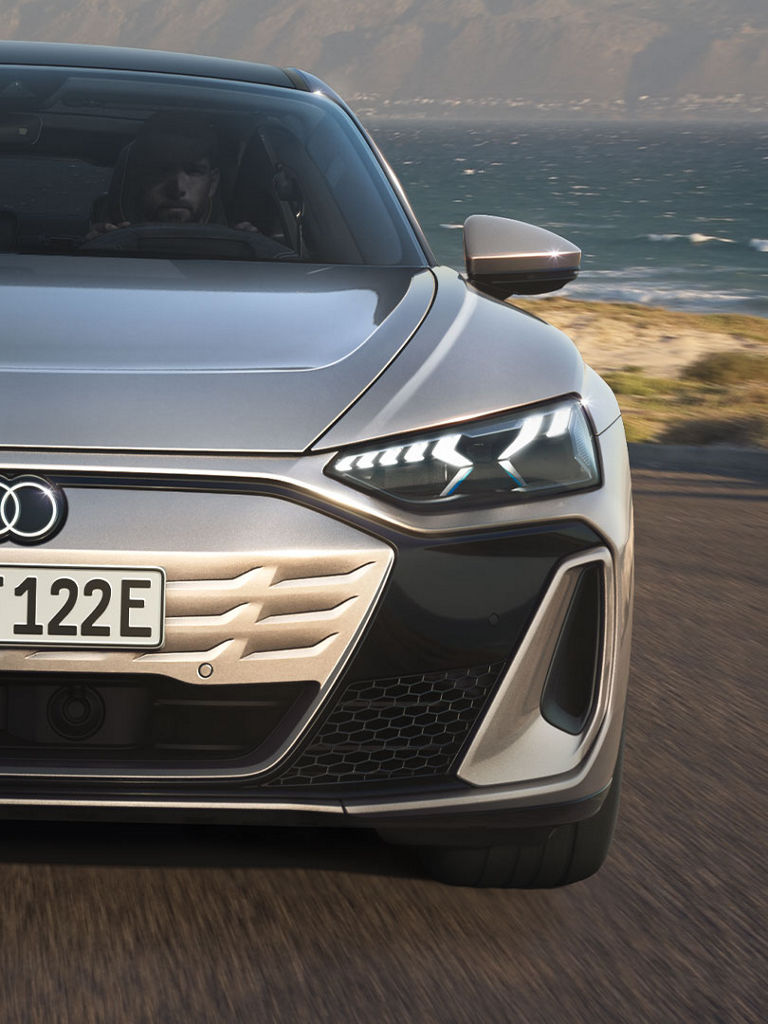 A frontal close-up of the Audi S e-tron GT on a coastal road. The focus is on the impressive light signature of the headlights and the sporty design.