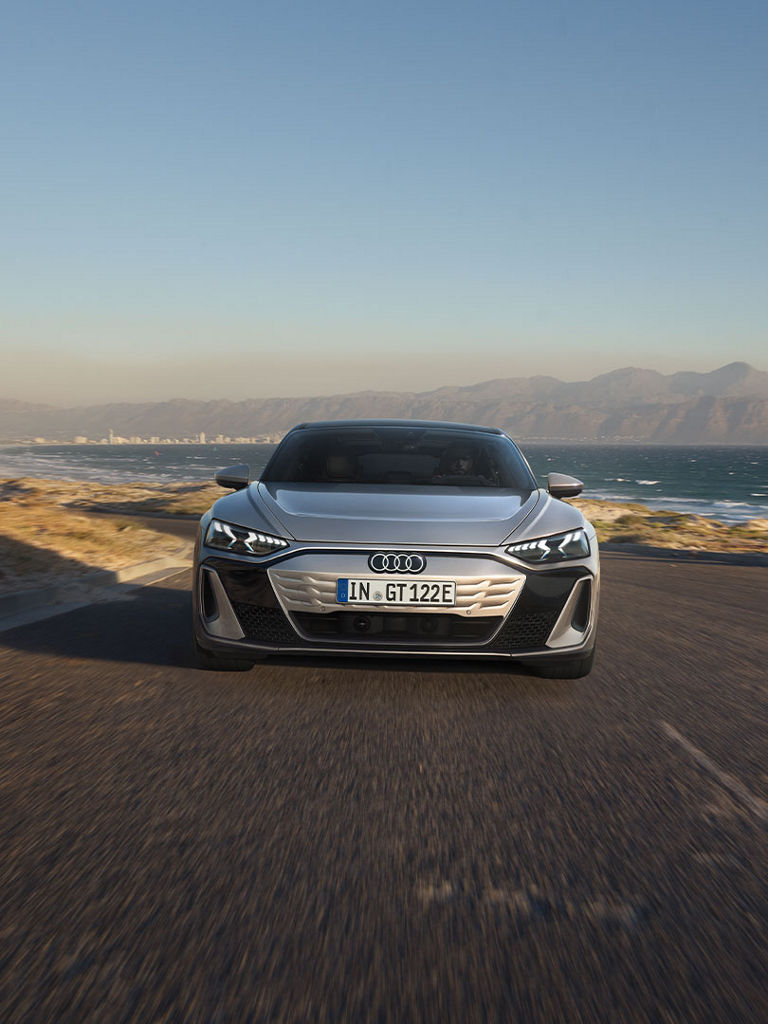  The front view of the Audi S e-tron GT on a winding coastal road. The striking LED headlights and the closed, aerodynamic radiator grille design are recognizable.