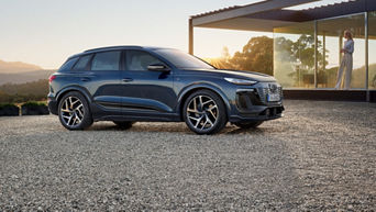  Dark blue Audi Q6 SUV e-tron standing on a gravel parking lot in front of a modern house, woman with smartphone in the background.